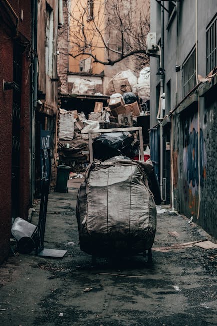 Two large black plastic bags filled with waste rest on a concrete sidewalk near a black metal fence, with a background of dense greenery and partially visible building structures. The bags appear to c